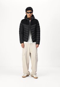 Chaqueta de plumas negra con capucha que presenta una sección superior texturizada, llevada sobre una camisa blanca, combinada con pantalones cargo beige y zapatillas deportivas.