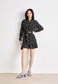 Abito nero con motivo floreale bianco e giallo, con bottoni sul davanti, maniche lunghe e orlo arricciato. Abbinato a tacchi blu con cinturini.