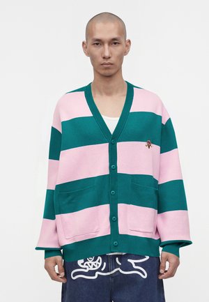 Mann trägt eine locker sitzende rosa-grün gestreifte Strickjacke mit aufgesetzten Taschen und Knopfverschluss, kombiniert mit dunklen Hosen mit weißem abstraktem Design.