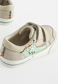 Beige sneakers med dobbelt Velcro-remme prydes af et tekstureret grønt og blåt broderet design på hælen, accentueret af små dekorative elementer.
