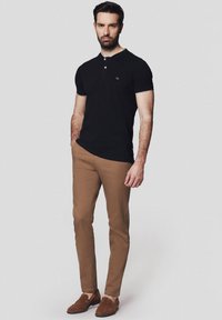 Czarna koszulka polo z dwoma guzikami, zestawiona z dopasowanymi beżowymi chinosami. Model nosi brązowe mokasyny, stojąc na neutralnym tle.