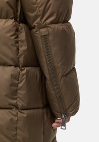 Braune Pufferjacke mit horizontalem Steppmuster, einem seitlichen Reißverschluss und einer weichen, glänzenden Oberfläche des Stoffes.