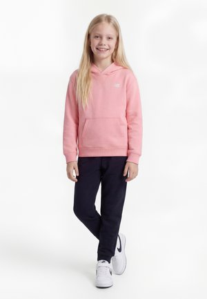 Fată tânără stând în picioare, purtând un hanorac roz, pantaloni negri și adidași albi, zâmbind pe un fundal alb simplu.