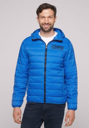 Man smiling wearing a blue quilted zip-up jacket with "CMPD Camp David" logo on the chest and dark jeans, standing against a plain background.