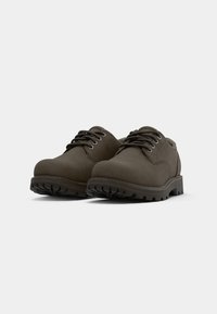 Ein Paar dunkelbraune Leder Schnürschuhe mit dicken Schlammsohlen, schräg von vorne betrachtet auf hellem, einfarbigen Hintergrund.