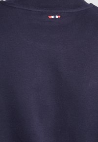 Tessuto di cotone blu navy con una consistenza liscia; cuciture di accento sottili in rosso, bianco e blu posizionate sul collo sul retro.