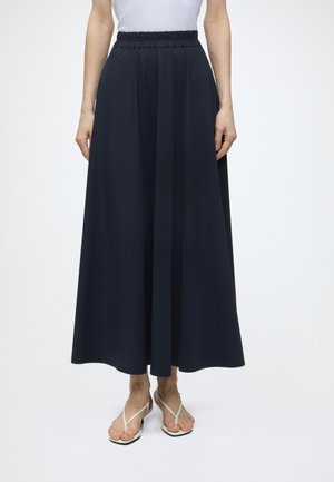 Femme portant une jupe longue taille haute bleu marine à jambes larges, associée à des sandales plates blanches à brides et un haut blanc.