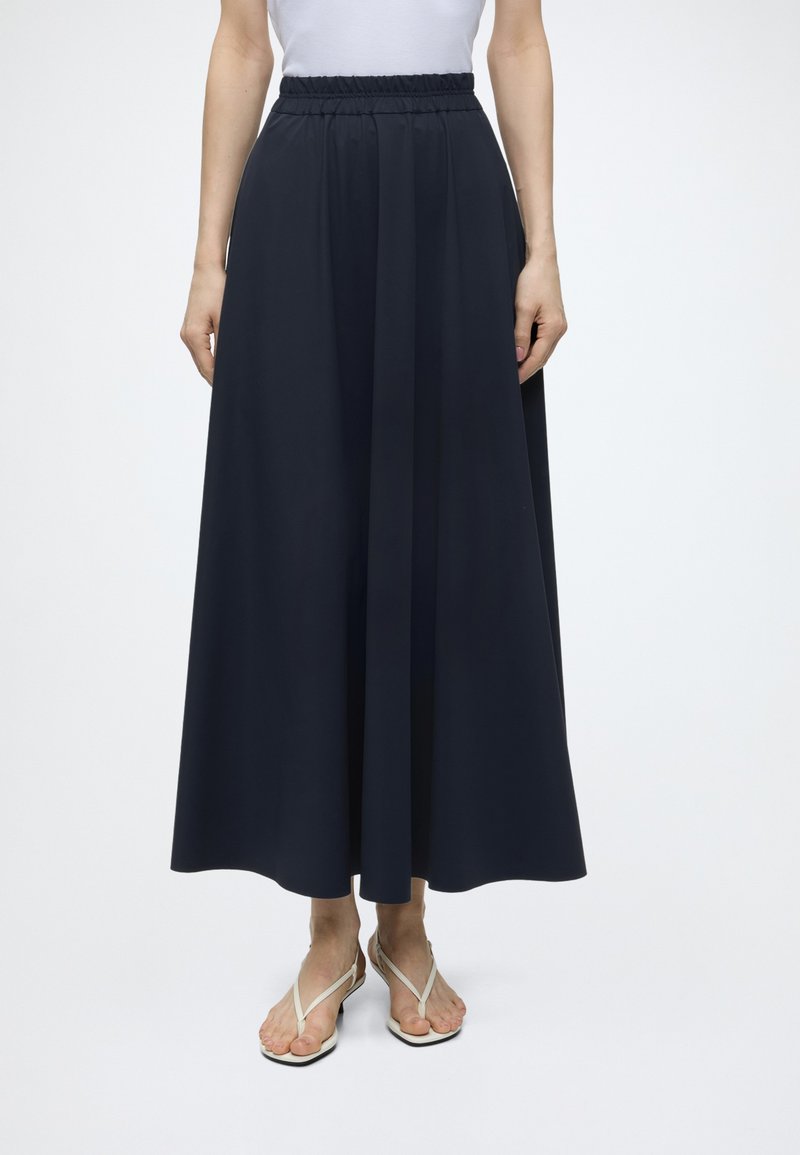 Femme portant une jupe longue taille haute bleu marine à jambes larges, associée à des sandales plates blanches à brides et un haut blanc.
