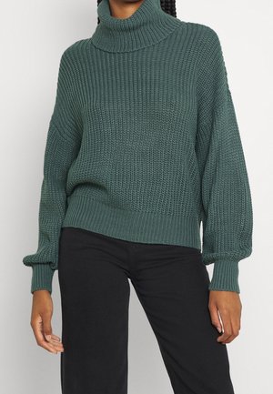 Grøn chunky strikket rullekravepullover med ballonsærmer båret sammen med sorte bukser på en person med fletninger.