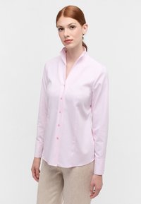 Camicia a maniche lunghe di colore rosa chiaro con tessuto strutturato, colletto e chiusura con bottoni. Presenta un orlo curvo e bottoni rosa a contrasto.