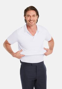 White polo shirt with a collar, short sleeves, and smooth texture, paired with dark trousers. Model is pulling the shirt upwards.