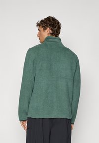Giacca in pile verde con colletto alto, tessuto texturizzato e vestibilità rilassata. La vista laterale mostra i dettagli delle cuciture e l'orlo.