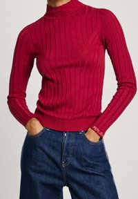 Gerippter Rollkragenpullover in lebhaftem Rot, aus weichem Stoff. Verfügt über lange Ärmel und dezentes Branding am Bündchen. Kombiniert mit blauen Jeans.