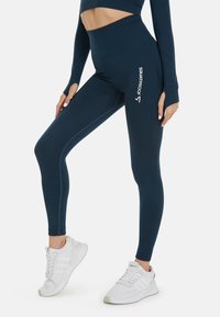 Tmavě modré legíny s vysokým pasem vyrobené z elastického materiálu; s logem "SQUATPROOF". Nosené s bílými teniskami.