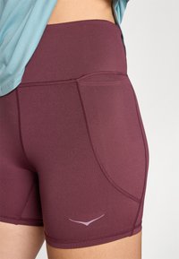 burgund rote Sportshorts mit hohem Bund, glatter Textur und lila Logo-Detail. Verfügt über eine Seitentasche und Nahtakzente.