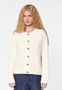 Cremefarbener Strickcardigan mit braunen Knöpfen, Rundhalsausschnitt und Rippbündchen. Das Design umfasst ein strukturiertes Quadratmuster, das sich über das gesamte Kleidungsstück erstreckt.