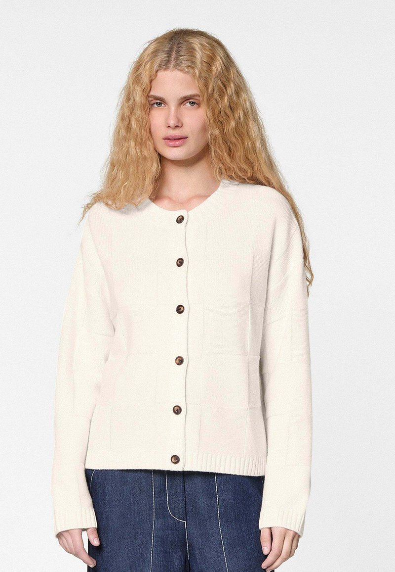 Cremefarbener Strickcardigan mit braunen Knöpfen, Rundhalsausschnitt und Rippbündchen. Das Design umfasst ein strukturiertes Quadratmuster, das sich über das gesamte Kleidungsstück erstreckt.