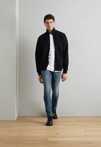Fiatal férfi sétál bent, fekete dzsekit, fehér inget, kék farmert és fekete bakancsot viselve, fapadon, sima világos falak mellett.