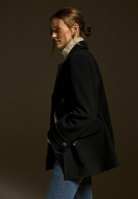 Svart dubbelknäppt kappa med rundade axlar, stora knappar och skräddarsydd passform, bärs över en cremefärgad polotröja och blå jeans.