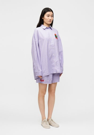 Γυναίκα που φορά υπερμεγέθη μωβ πουκάμισο καρό με κεντήματα λουλουδιών και αντίστοιχα σορτς με κόκκινη κεντημένη γραφή, μπεζ αθλητικά παπούτσια.