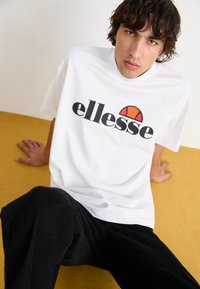 Valge lühikeste varrukatega T-särk, millel on must "ellesse" logo ja punane-oranž poolringikujuline disain. Kombineeritud mustade tekstureeritud pükstega.