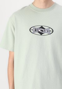 Light green cotton t-shirt featuring a black and blue Rip Curl logo in an oval design on the chest. Crew neck with short sleeves.