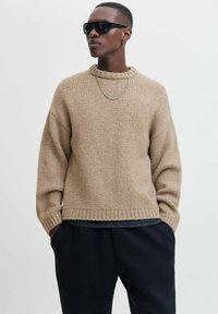 Beige gestrickter Pullover mit hohem Rippenkragen und weiten Ärmeln. Über einem schwarzen Oberteil getragen, kombiniert mit schwarzen Hosen.