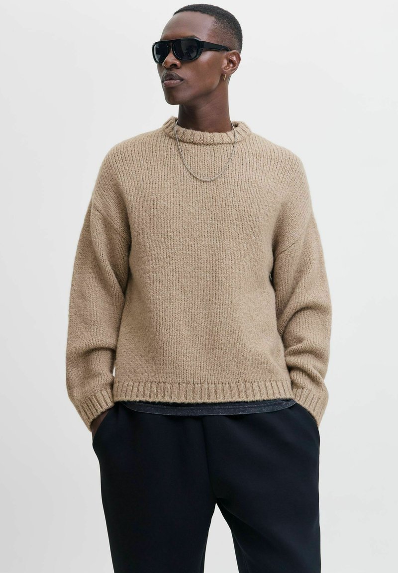Beige gestrickter Pullover mit hohem Rippenkragen und weiten Ärmeln. Über einem schwarzen Oberteil getragen, kombiniert mit schwarzen Hosen.