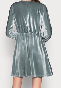 Vestido de terciopelo en verde azulado claro con mangas largas, cintura ajustada y falda acampanada. Textura suave y brillo sutil.