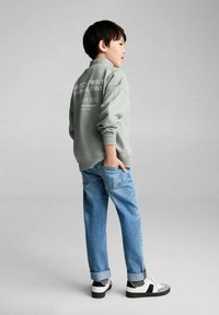 Grijze sweatshirt met rugafdruk tekst, losse pasvorm. Lichtblauwe denim jeans met opgerolde boorden. Zwart-witte sneakers. Achtergrond is lichtgrijs.