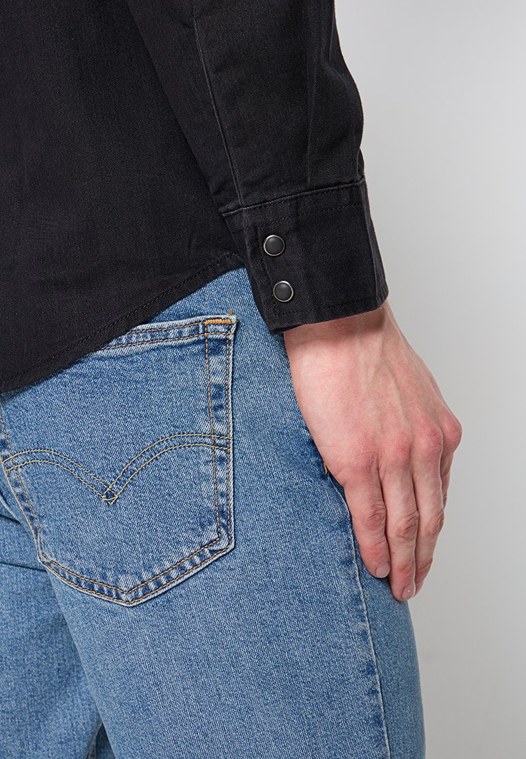 Camicia di jeans nera con polsini abbottonati, abbinata a jeans blu con tasche posteriori distintive e un motivo di cucitura a contrasto.