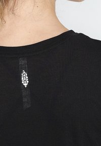 Musta ribattu t-paita, jossa on pyöreä pääntie ja pieni valkoinen kukkakuosi selkäpuolella, joka korostaa kankaan rakennetta.