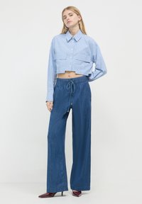 Frau steht und trägt ein kurzes blau gestreiftes Hemd, weit geschnittene blaue Hose mit Kordelzug und burgunderfarbene spitze High Heels.