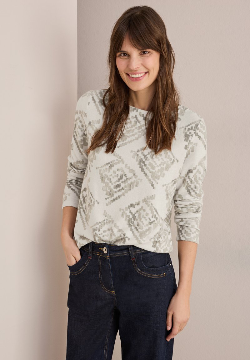 Hellgrauer Langarmpullover mit einem geometrischen Muster in dunklerem Grau, kombiniert mit dunkelblauen Jeans mit orangefarbenen Nähten.