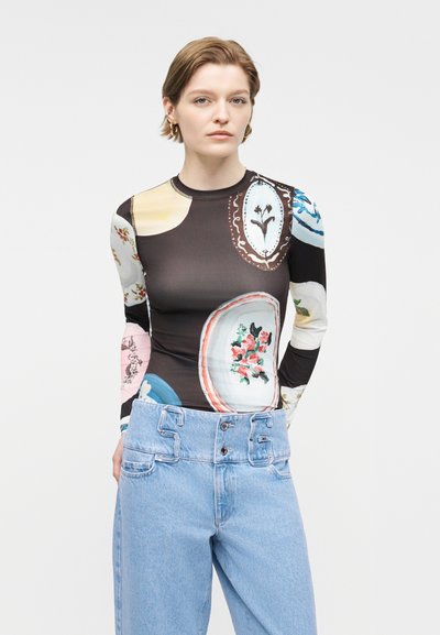 Γυναίκα με κοντά μαλλιά φοράει εφαρμοστή μπλούζα με μακριά μανίκια και πολύχρωμα λουλουδένια prints, καθώς και ψηλόμεσο ανοιχτό μπλε τζιν παντελόνι.