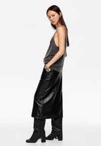 Top de tirantes de punto gris, falda maxi de cuero negra, botas hasta la rodilla a juego y pulsera de plata. La modelo posa de lado contra un fondo blanco.