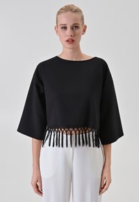Top nero accorciato con maniche ampie, caratterizzato da un dettaglio di frange sull'orlo. Il tessuto appare liscio con una silhouette strutturata.
