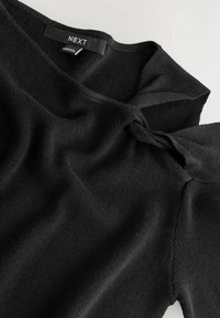 Pull en maille noire avec un détail torsadé près de l'encolure et une étiquette de marque visible portant l'inscription "NEXT" à l'intérieur du col.
