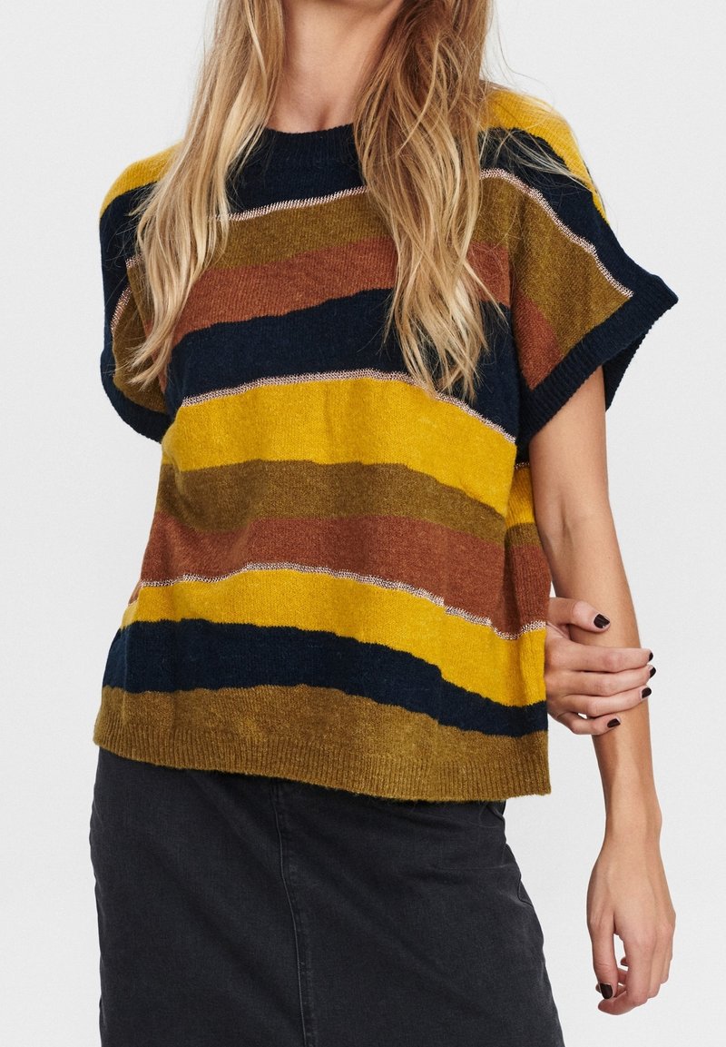 Femme portant un pull ample à manches courtes avec de larges rayures jaunes, brunes et bleu marine, et une jupe sombre, debout devant un fond uni.