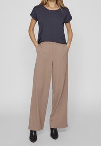 Žena v námořnickém krátkém rukávu, béžových širokých kalhotách a černých botách s špičatou špičkou, stojí s rukama v kapsách.