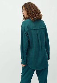Maglietta a maniche lunghe color teal con taglio dritto, polsini con bottoni e dettaglio di pieghe sul retro. Realizzata in un tessuto testurizzato e morbido.