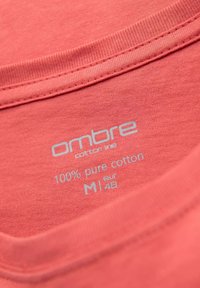 Korálové bavlněné tričko s etiketou, na které je šedým písmem napsáno: "ombre bavlněná řada, 100 % čistá bavlna, M, eur 48." Hladký výstřih a měkká textura.