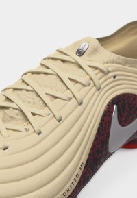 Κοντινή λήψη σε μπεζ αθλητικό παπούτσι με υφασμάτινο επάνω μέρος με υφή, κορδόνια που διασταυρώνονται, κόκκινο και μαύρο ένθετο σόλας με σχέδιο και μεγάλο γκρι λογότυπο Nike swoosh.