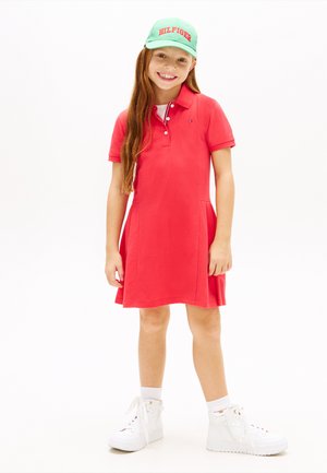 Ragazza sorridente con lunghi capelli rossi che indossa un vestito polo rosso, sneakers alte bianche, calzini bianchi e un cappellino verde con lettere rosse.