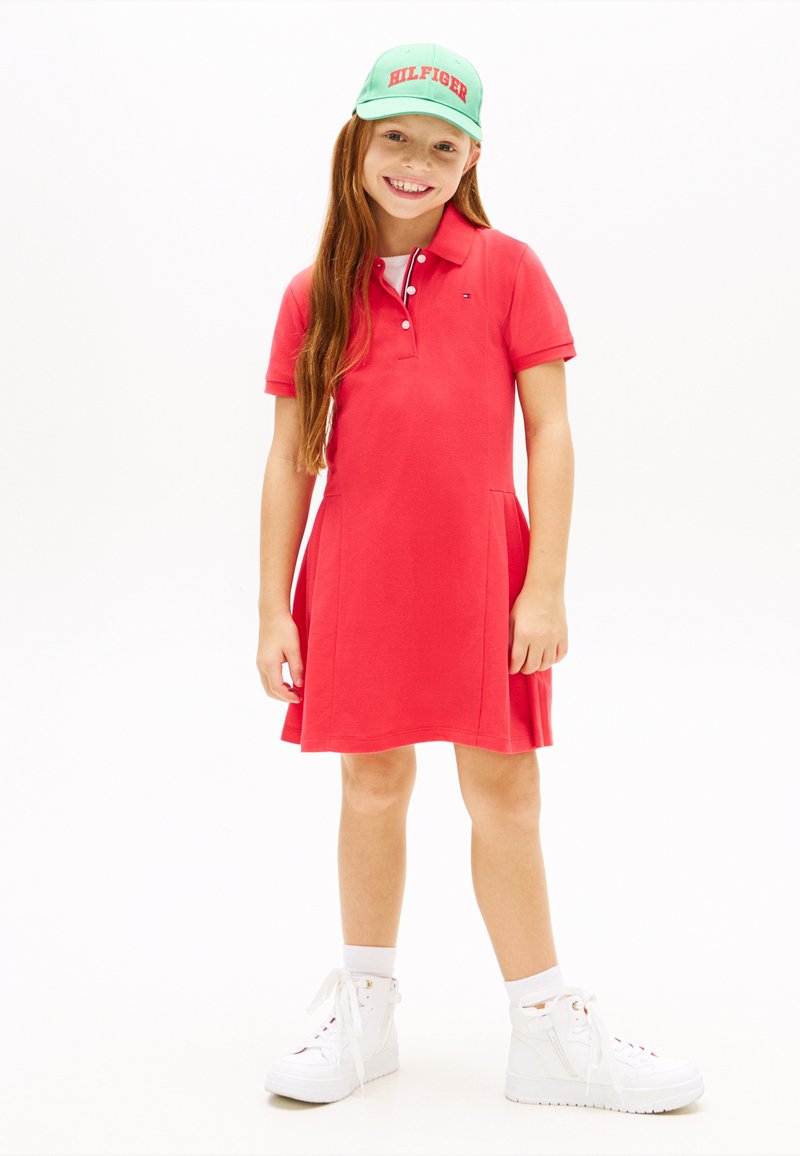 Χαμογελαστή κοπέλα με μακριά κόκκινα μαλλιά, ντυμένη με κόκκινο φόρεμα πόλο, λευκά ψηλά αθλητικά παπούτσια, λευκές κάλτσες και πράσινο καπέλο με κόκκινα γράμματα.