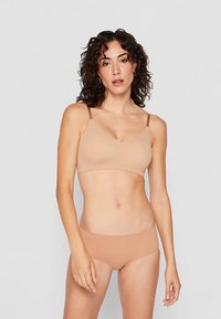 Mladá žena s kučeravými vlasmi, oblečená v bezšvovej telovej podprsenke a zodpovedajúcich nohavičkách, stojaca na jednoduchom svetlošedom pozadí.