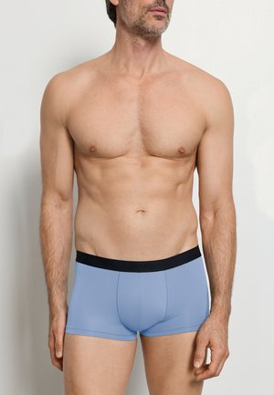 Mężczyzna bez koszulki, ubrany w jasnoniebieskie bokserki z czarnym paskiem w pasie, stojący na jasnym, jednolitym tle, lekko zwrócony w prawo.