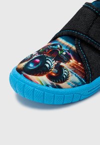 Zapato de tela negra con un gráfico vibrante de un monster truck, con suela de goma azul con agarre texturizado y correa ajustable.