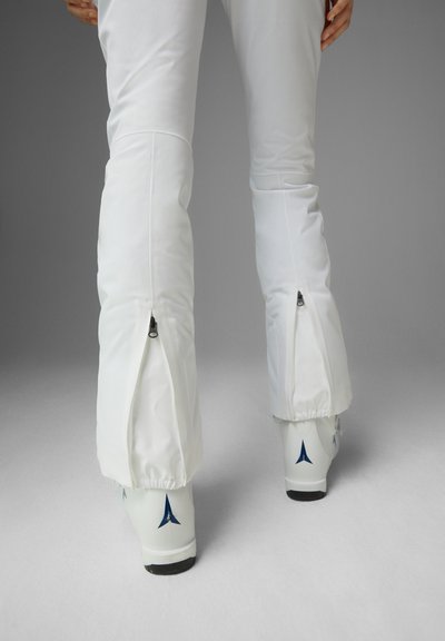 Weiße Skihosen mit Reißverschlüssen an den Knöcheln. Das Material wirkt glatt, und die Hose ist mit weißen Skischuhen mit blauen Akzenten kombiniert.