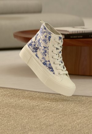 Basket montante avec motif floral bleu et papillons sur tissu blanc, semelle blanche épaisse, placée à l'intérieur près d'une table en bois et d'un canapé.